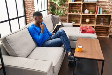 Young african american man reading book sitting on sofa at home