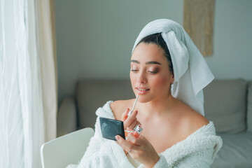 Young beautiful woman in terry white dressing gown makes herself beautiful makeup at home. Model holds makeup brush and cosmetics in her hands.