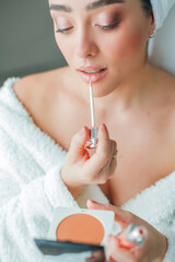 Young beautiful woman in terry white dressing gown makes herself beautiful makeup at home. Model holds makeup brush and cosmetics in her hands.