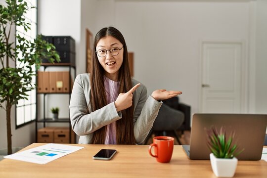 Young chinese business worker wearing business style sitting on desk at office amazed and smiling to the camera while presenting with hand and pointing with finger.