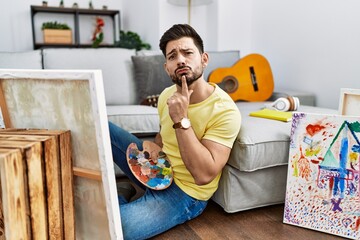 Young man with beard painting canvas at home thinking concentrated about doubt with finger on chin...