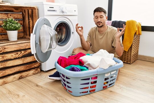 Young handsome man putting dirty laundry into washing machine relax and smiling with eyes closed doing meditation gesture with fingers. yoga concept.