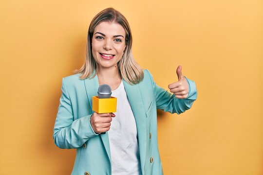 Beautiful caucasian woman holding reporter microphone smiling happy and positive, thumb up doing excellent and approval sign
