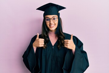 Young hispanic woman wearing graduation cap and ceremony robe success sign doing positive gesture...