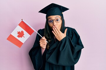 Young hispanic woman wearing graduation uniform holding canada flag covering mouth with hand, shocked and afraid for mistake. surprised expression