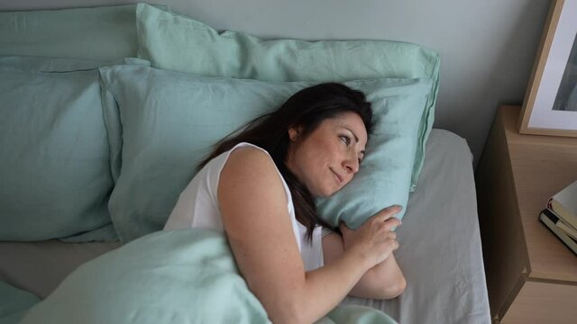 Top view portrait of a smiling woman in a comfortable bed with blue linen sheets, pensive and happy resting, waking up in the morning after a good sleep in a cozy home or hotel room