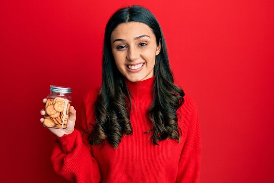 Young hispanic woman holding salty biscuits jar looking positive and happy standing and smiling with a confident smile showing teeth