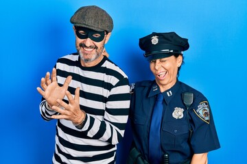 Middle age couple of hispanic woman and man wearing thief and police uniform winking looking at the...