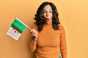 Middle age hispanic woman holding algeria flag scared and amazed with open mouth for surprise, disbelief face