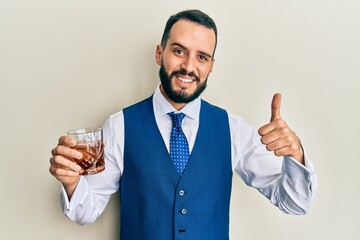 Young man with beard drinking whiskey shot smiling happy and positive, thumb up doing excellent and approval sign