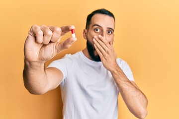 Young man with beard holding pill covering mouth with hand, shocked and afraid for mistake....