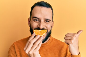 Young man with beard holding orange slice on mouth as funny smile pointing thumb up to the side...