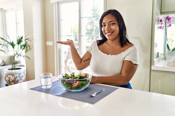 Obraz na płótnie Canvas Young hispanic woman eating healthy salad at home smiling cheerful presenting and pointing with palm of hand looking at the camera.