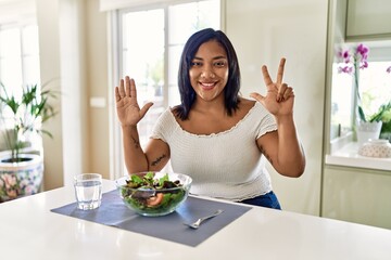 Obraz na płótnie Canvas Young hispanic woman eating healthy salad at home showing and pointing up with fingers number eight while smiling confident and happy.