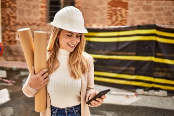Young blonde woman holding blueprints using smartphone at street