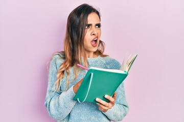 Young hispanic girl reading and writing book angry and mad screaming frustrated and furious, shouting with anger. rage and aggressive concept.