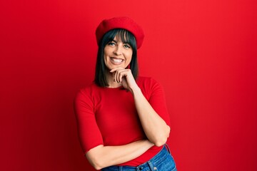 Naklejka premium Young hispanic woman wearing french look with beret looking confident at the camera with smile with crossed arms and hand raised on chin. thinking positive.