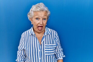 Senior woman with grey hair standing over blue background sticking tongue out happy with funny expression. emotion concept.