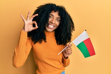 African american woman with afro hair holding madagascar flag doing ok sign with fingers, smiling friendly gesturing excellent symbol