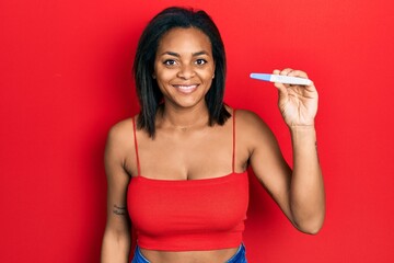 Young african american girl holding pregnancy test result looking positive and happy standing and smiling with a confident smile showing teeth