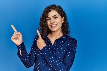 Young brunette woman with curly hair wearing casual clothes over blue background smiling and looking at the camera pointing with two hands and fingers to the side.