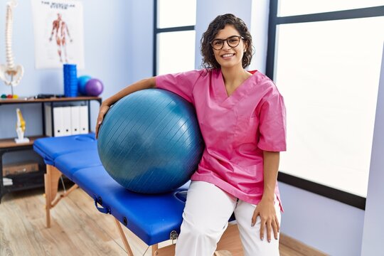 Young latin woman wearing physiotherapist uniform holding fit ball at clinic