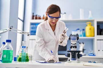 Middle age woman wearing scientist uniform writing on clipboard holding test tubes at laboratory