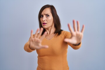 Middle age brunette woman standing wearing orange sweater afraid and terrified with fear expression stop gesture with hands, shouting in shock. panic concept.