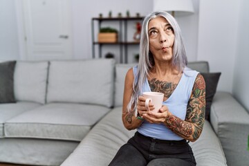 Middle age grey-haired woman drinking coffee sitting on the sofa at home making fish face with...