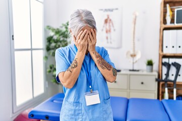 Middle age grey-haired woman wearing physiotherapist uniform at medical clinic with sad expression covering face with hands while crying. depression concept.