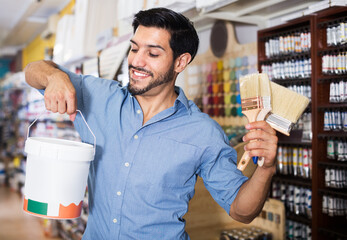 Smiling male buyer holding bucket of paint and brushes in paint shop