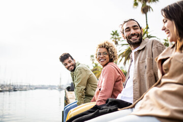 Young group of diverse adult friends laughing while sitting in front of the sea in Barcelona - United multi ethnic people enjoying together outdoors - obrazy, fototapety, plakaty