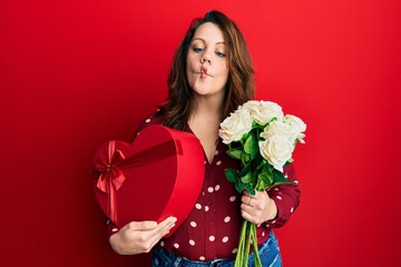 Young caucasian woman holding valentine gift and flowers making fish face with mouth and squinting eyes, crazy and comical.