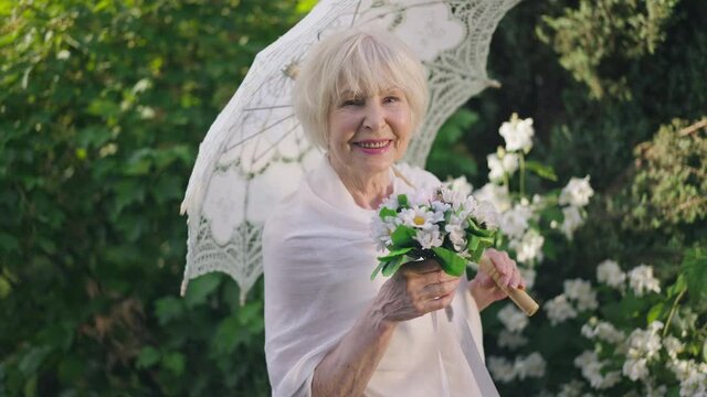 Elegant senior bride with bouquet and sun umbrella posing in slow motion on sunny spring summer day in park. Portrait of beautiful happy Caucasian woman with grey hair looking at camera smiling