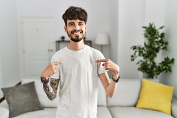 Hispanic man with beard at the living room at home looking confident with smile on face, pointing...