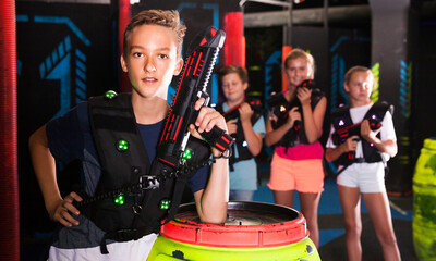 Obraz na płótnie Canvas Portrait of teen boy standing with laser gun on dark lasertag arena on background with his team..