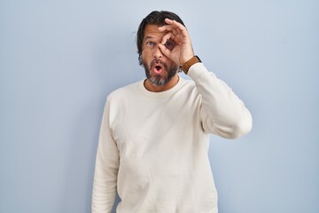 Handsome middle age man wearing casual sweater over blue background doing ok gesture shocked with surprised face, eye looking through fingers. unbelieving expression.