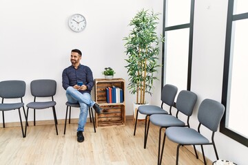 Young hispanic man using smartphone sitting on chair at waiting room