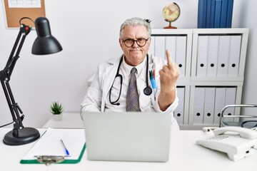Senior caucasian man wearing doctor uniform and stethoscope at the clinic showing middle finger, impolite and rude fuck off expression