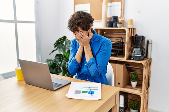 Young hispanic man with beard working at the office using computer laptop with sad expression covering face with hands while crying. depression concept.