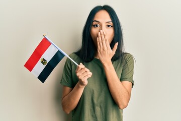 Young hispanic girl holding egypt flag covering mouth with hand, shocked and afraid for mistake....