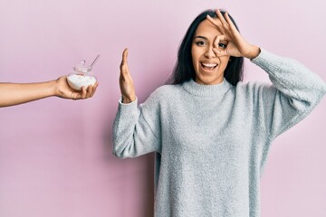 Young hispanic girl rejecting bowl with sugar smiling happy doing ok sign with hand on eye looking...