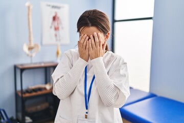 Young brunette woman working at pain recovery clinic with sad expression covering face with hands while crying. depression concept.