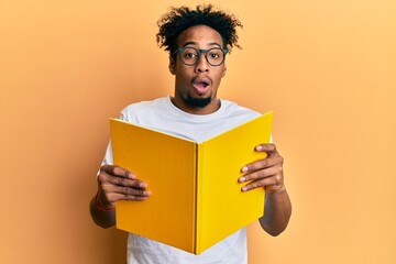 Young african american man with beard reading a book wearing glasses afraid and shocked with...