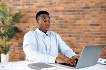 Get unexpected news. African american black shocked man, office employee, CEO, looking in surprise at the laptop screen on modern office, received an unexpected message, amazed facial expression