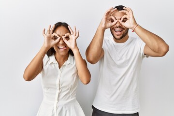 Young interracial couple standing together in love over isolated background doing ok gesture like binoculars sticking tongue out, eyes looking through fingers. crazy expression.