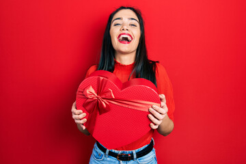 Young hispanic girl holding valentine gift smiling and laughing hard out loud because funny crazy...