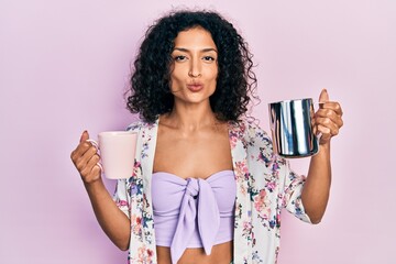 Young latin girl holding cup of coffee and jar of milk looking at the camera blowing a kiss being...