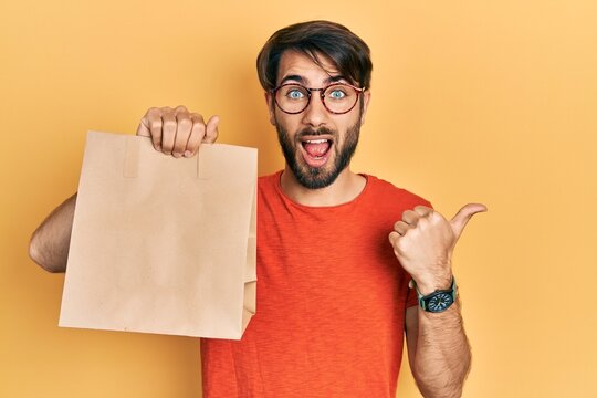 Young hispanic man holding paper bag with bread pointing thumb up to the side smiling happy with open mouth