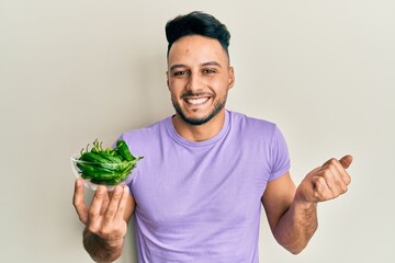 Young arab man holding bowl of green peppers screaming proud, celebrating victory and success very...